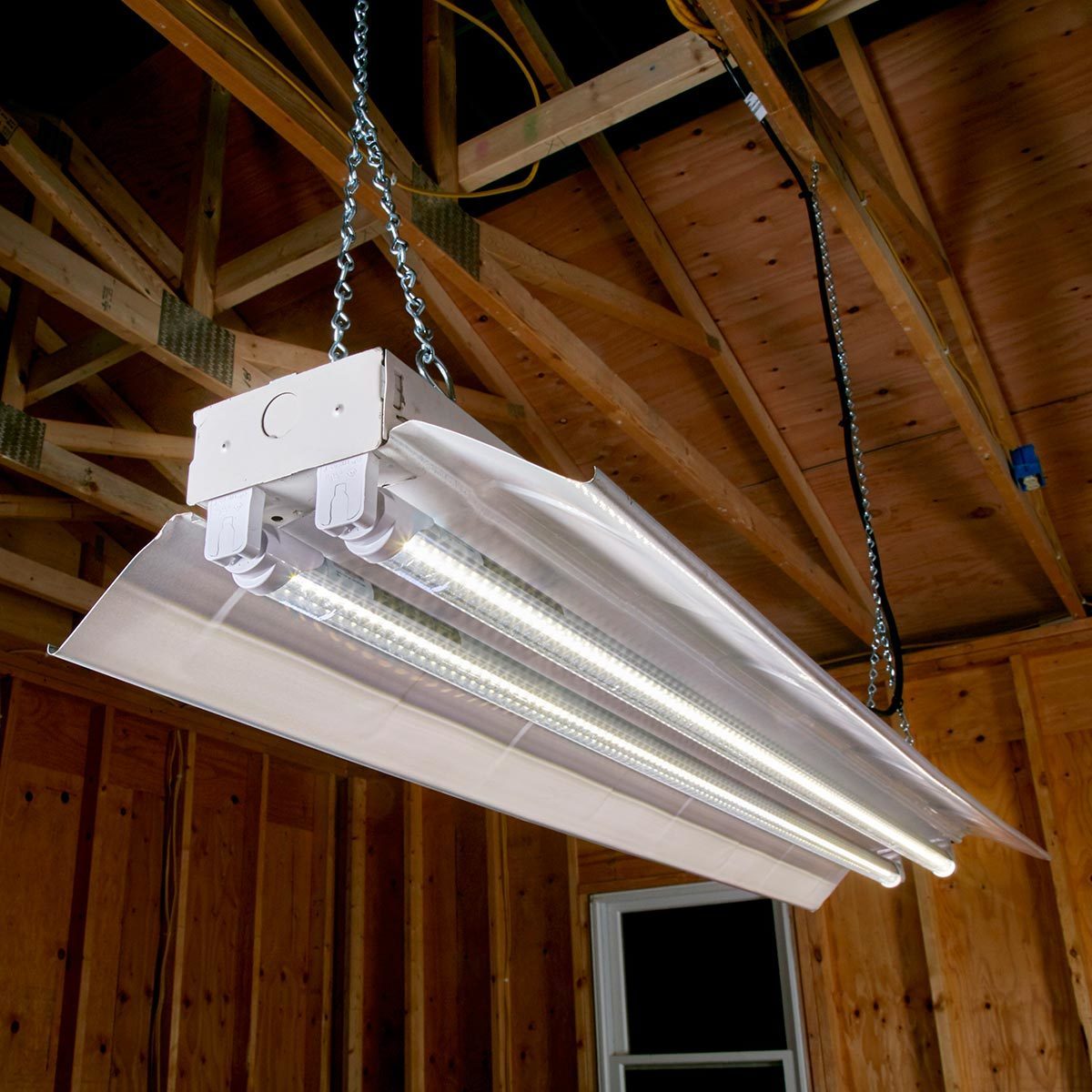 A fluorescent light fixture hangs from the ceiling, illuminating a wooden space with exposed beams and a window, creating a functional workspace atmosphere.