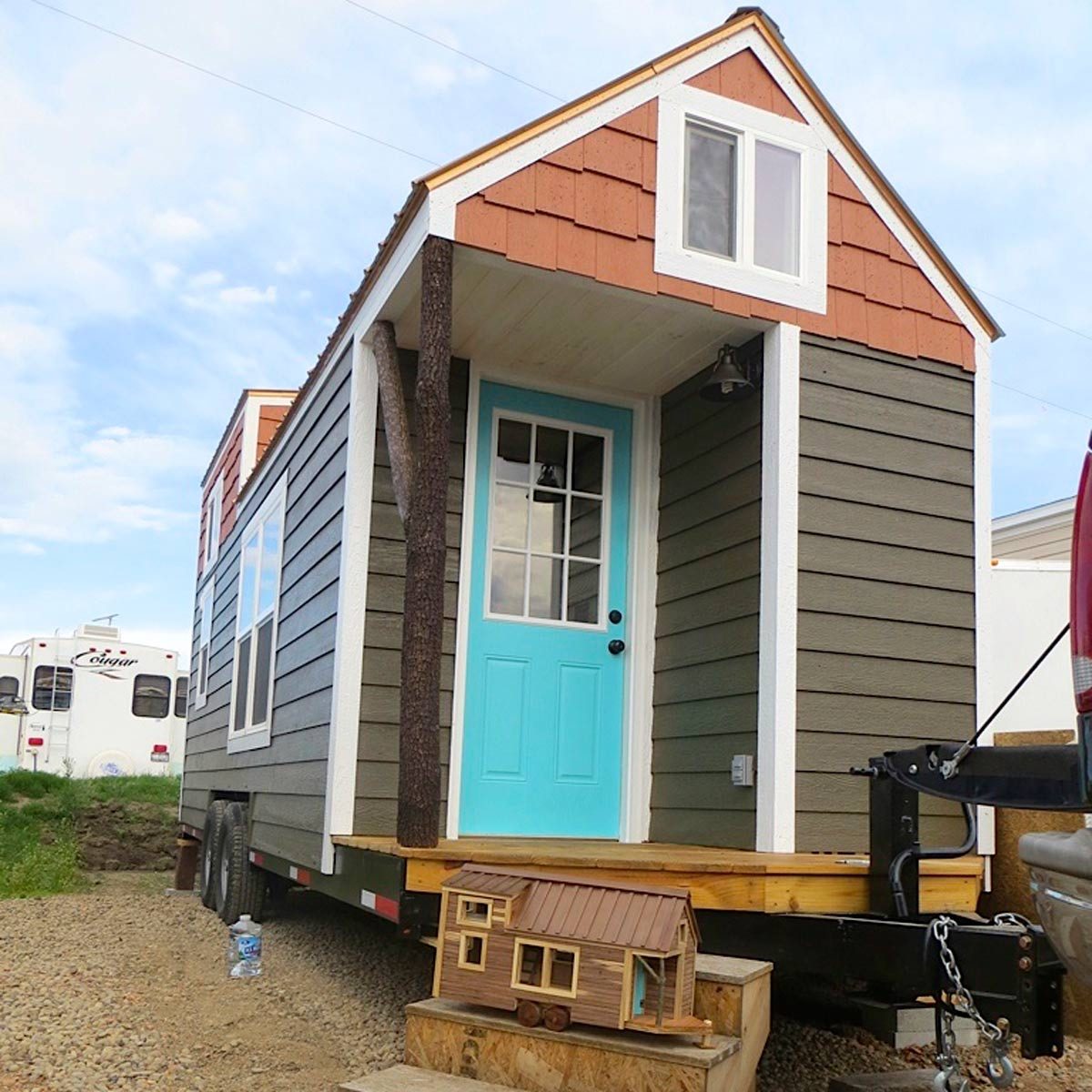 A tiny house with a turquoise door sits on a trailer, featuring wooden shingles and a porch, surrounded by gravel and a blue sky.