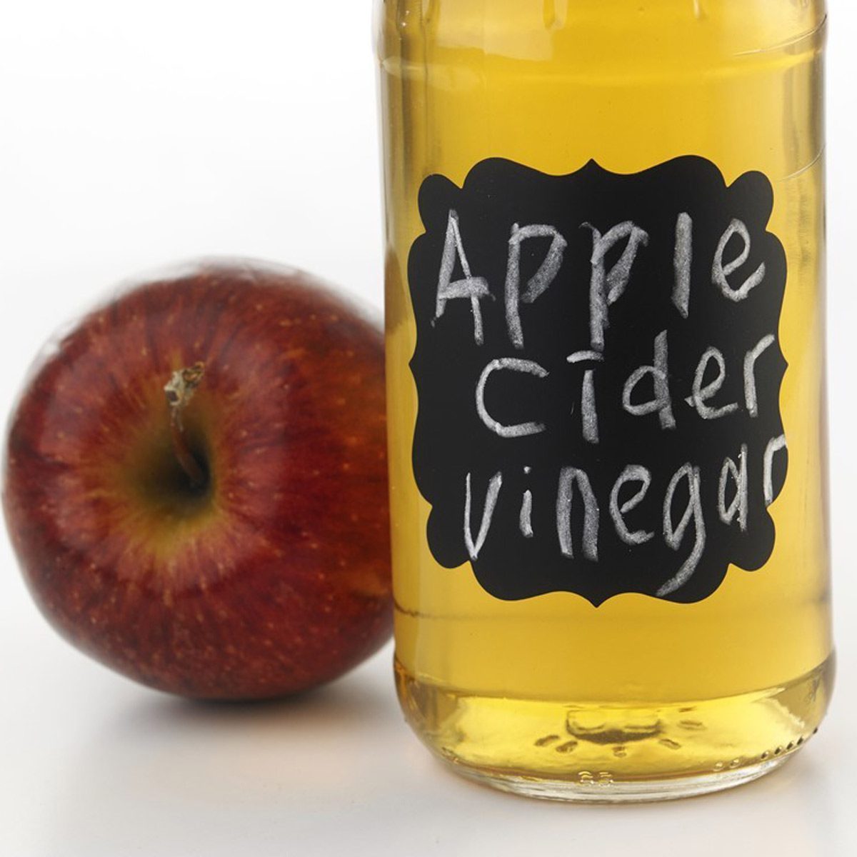A bottle of apple cider vinegar stands next to a red apple on a white surface, labeled with handwritten text.