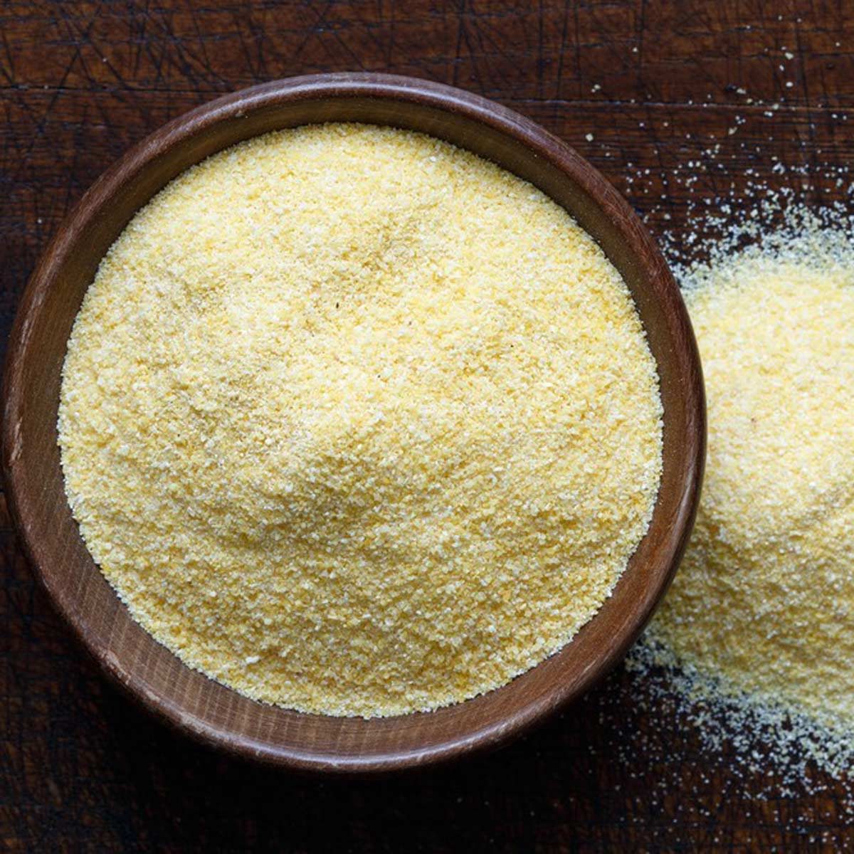 A wooden bowl contains yellow cornmeal, with some spilled onto a dark, textured surface. The fine grains appear dry and granular in texture.