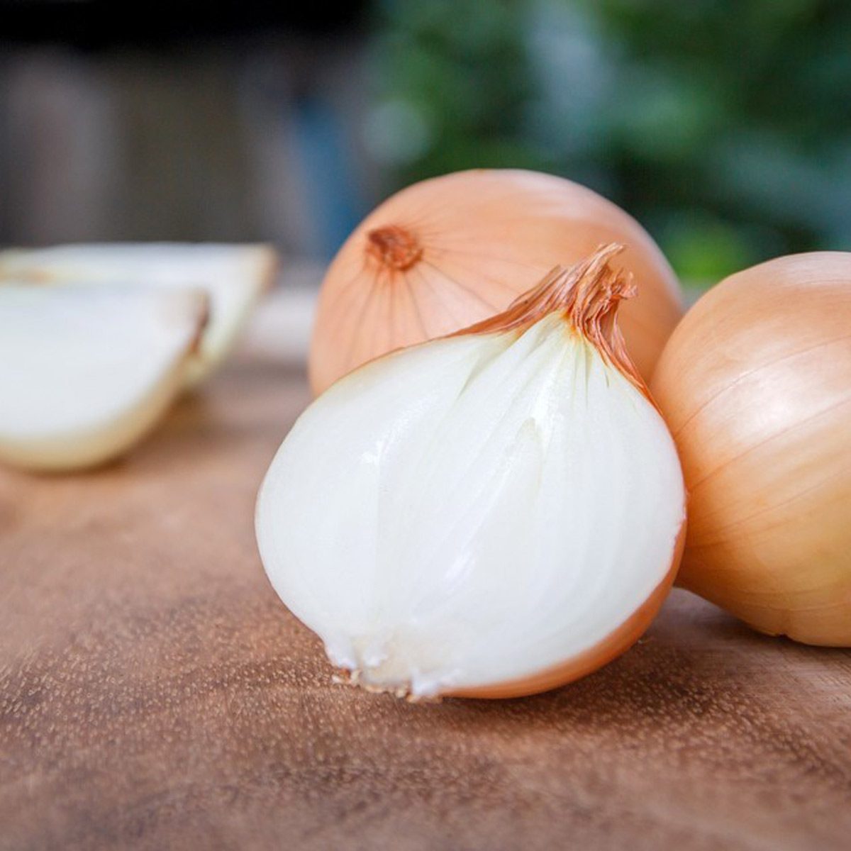 Sliced onion reveals its white interior, resting on a wooden surface with whole onions nearby, set against a blurred green background.