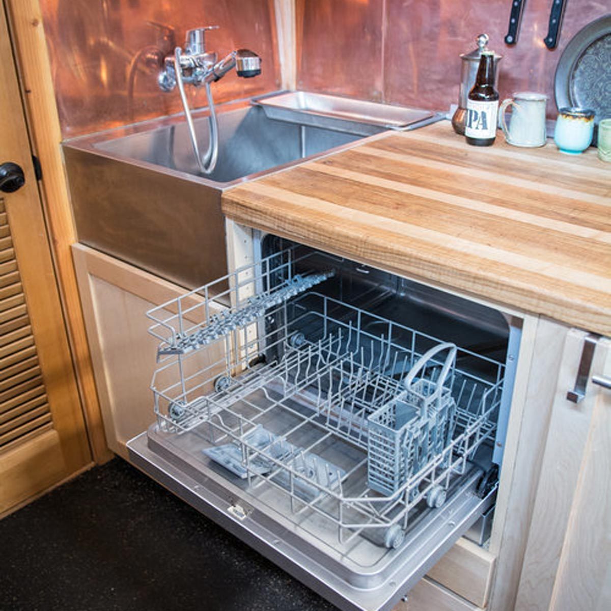 A dishwasher stands open, revealing its interior. Nearby, a stainless steel sink with a faucet and a wooden countertop are visible in a kitchen setting.