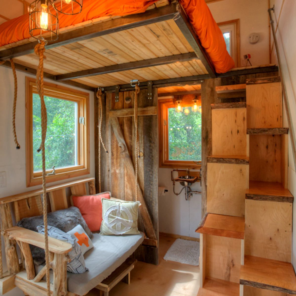 A lofted bed hangs above a cozy sitting area with rustic wooden furniture, illuminated by unique pendant lights, surrounded by windows featuring a green outdoor view.