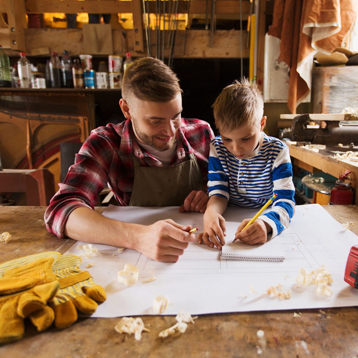 A man and a boy draw together on a large sheet of paper in a workshop, surrounded by tools and wood shavings.