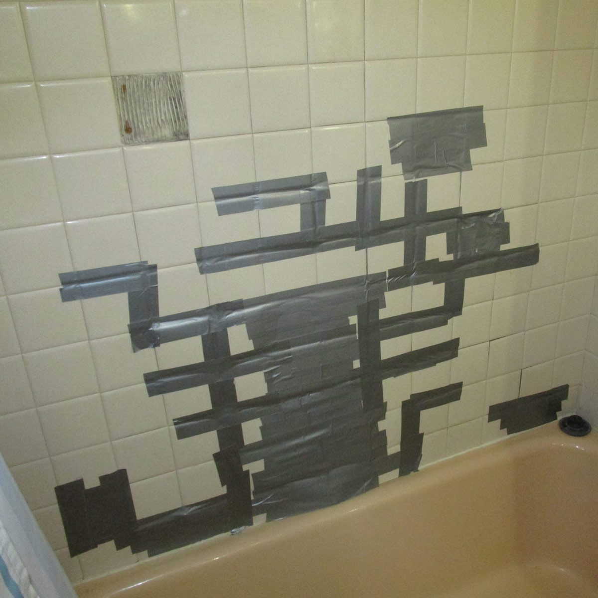 A section of tiled bathroom wall is covered with overlapping strips of gray tape, concealing a small missing tile above a bathtub.