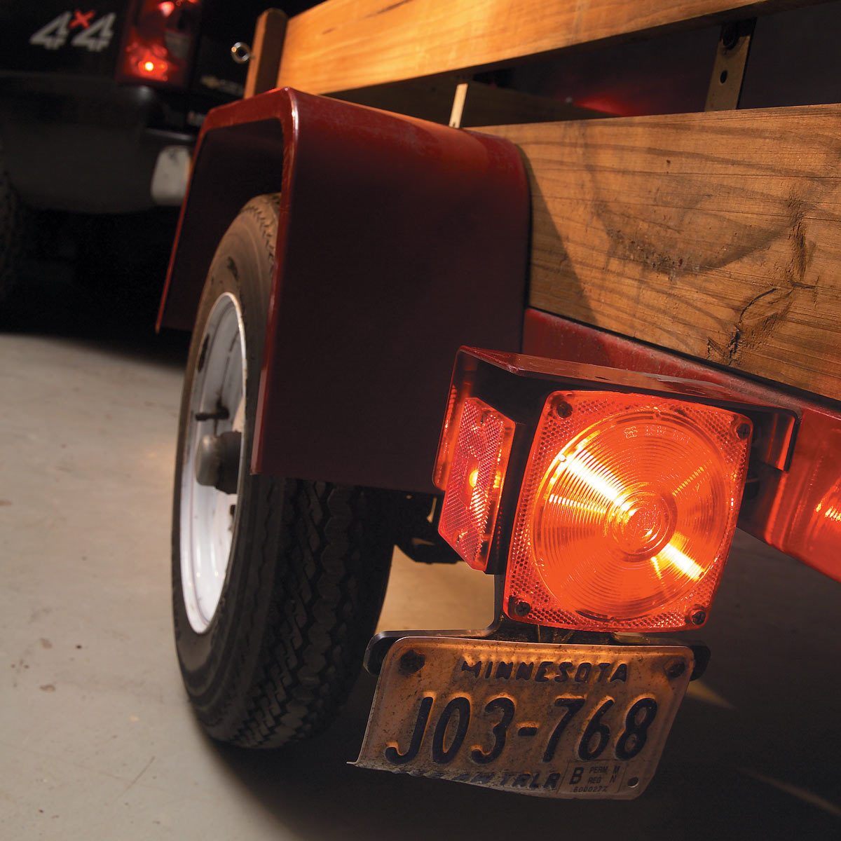 A trailer's rear shows a red light flashing, resting in a dimly lit garage with part of a vehicle visible in the background.