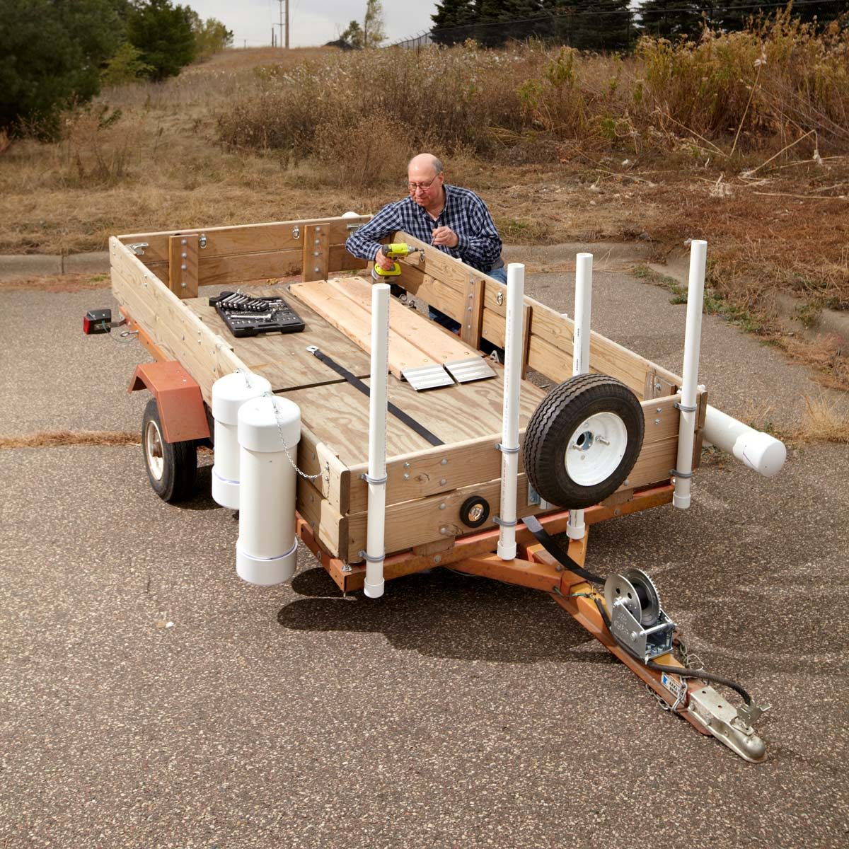 A man is working on a wooden trailer, using a measuring tool, on a gravel surface surrounded by sparse vegetation and unpaved terrain.