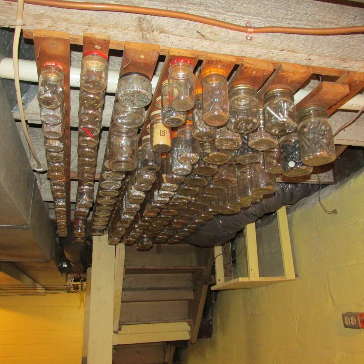 Glass jars are suspended from wooden beams, containing various small items. The setting is a basement with exposed pipes and yellow-painted walls.