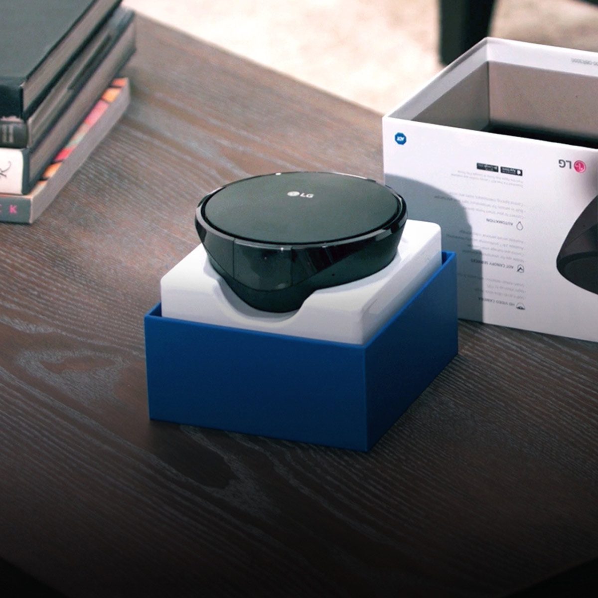 A black electronic device sits on a white cushion within a blue box, surrounded by books and an open product packaging on a wooden table.