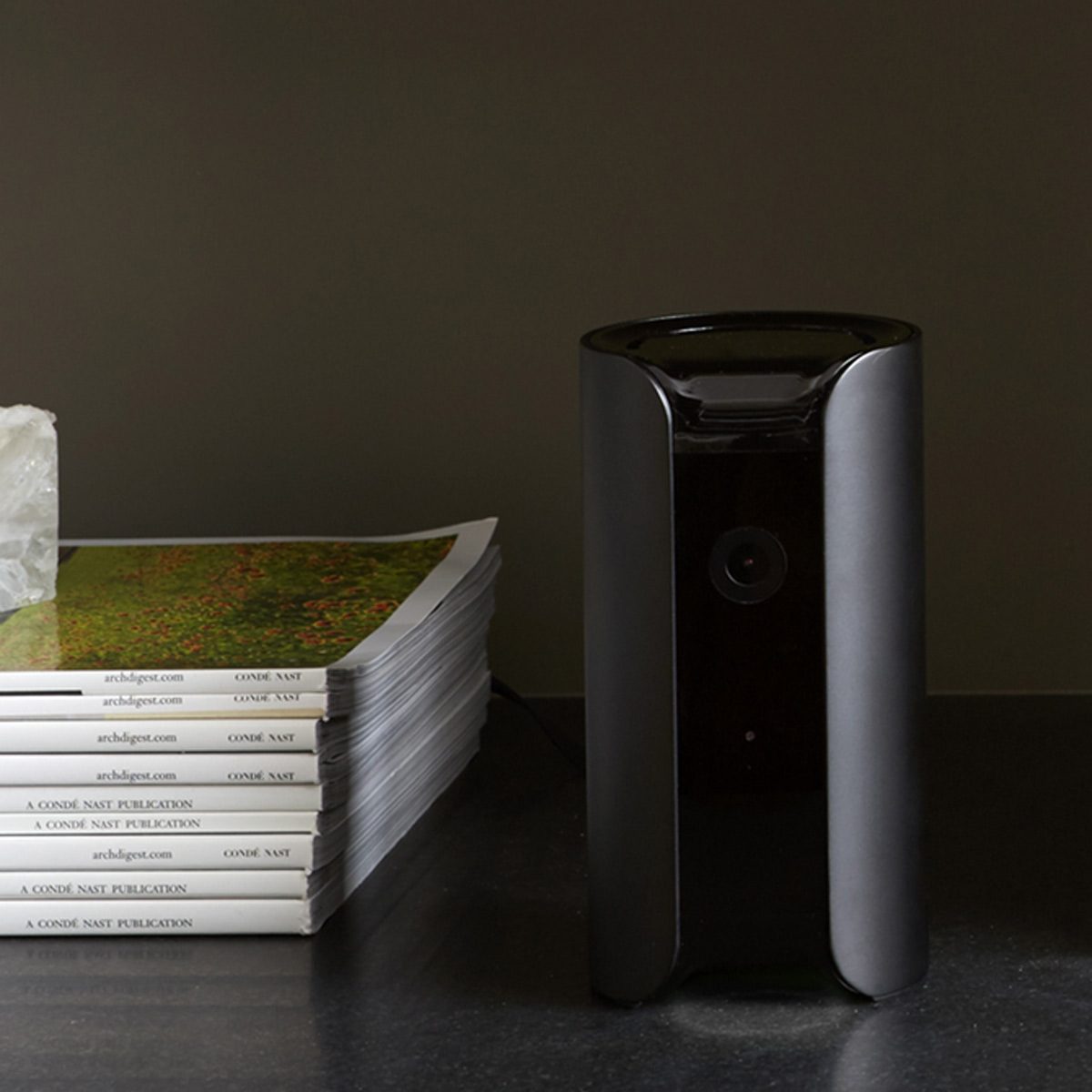 A sleek black device stands beside a stack of magazines on a dark surface, with a translucent crystal nearby, in a minimalistic indoor setting.