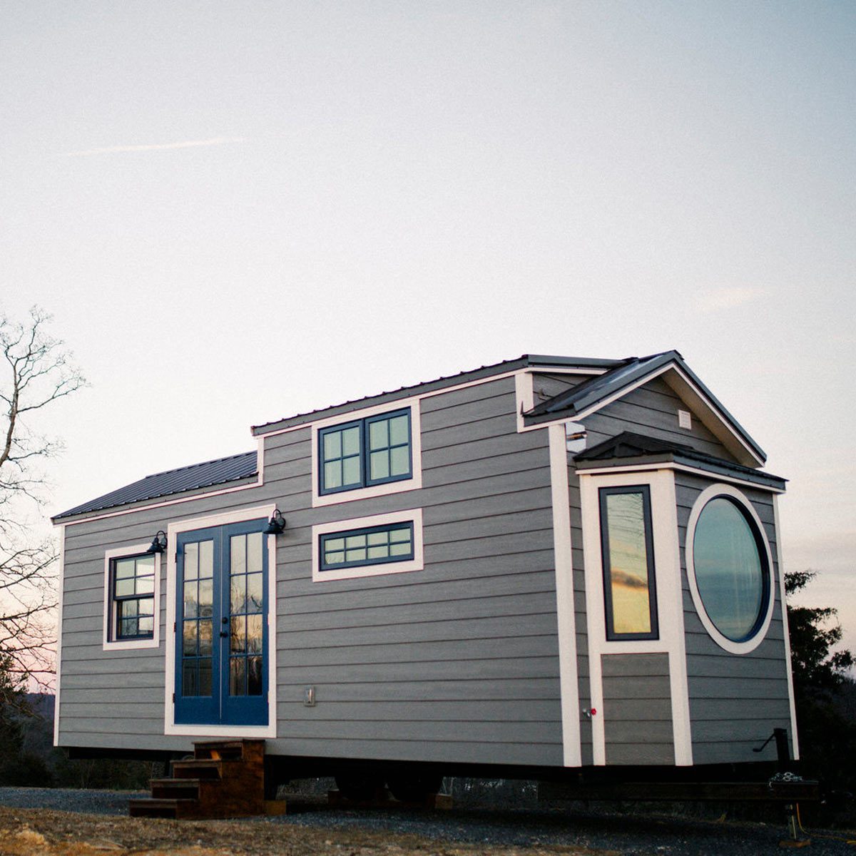 A small, modern house with a blue front door sits on wheels, surrounded by bare trees under a pastel sky at dusk.