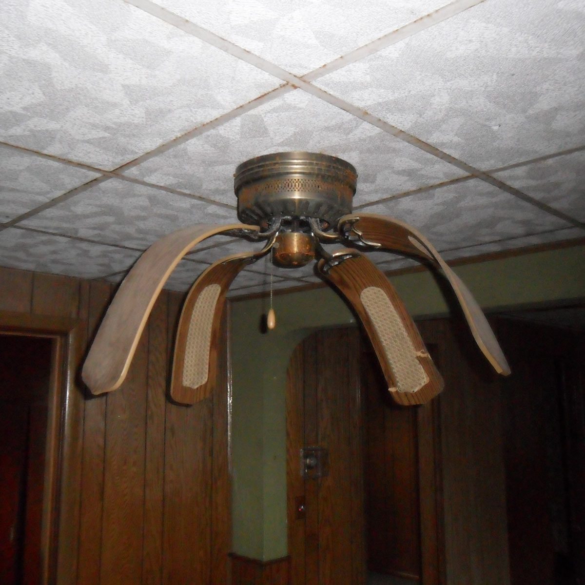A ceiling fan with five wooden blades spins slowly, hanging from a textured ceiling in a room with wooden paneling and doors in the background.
