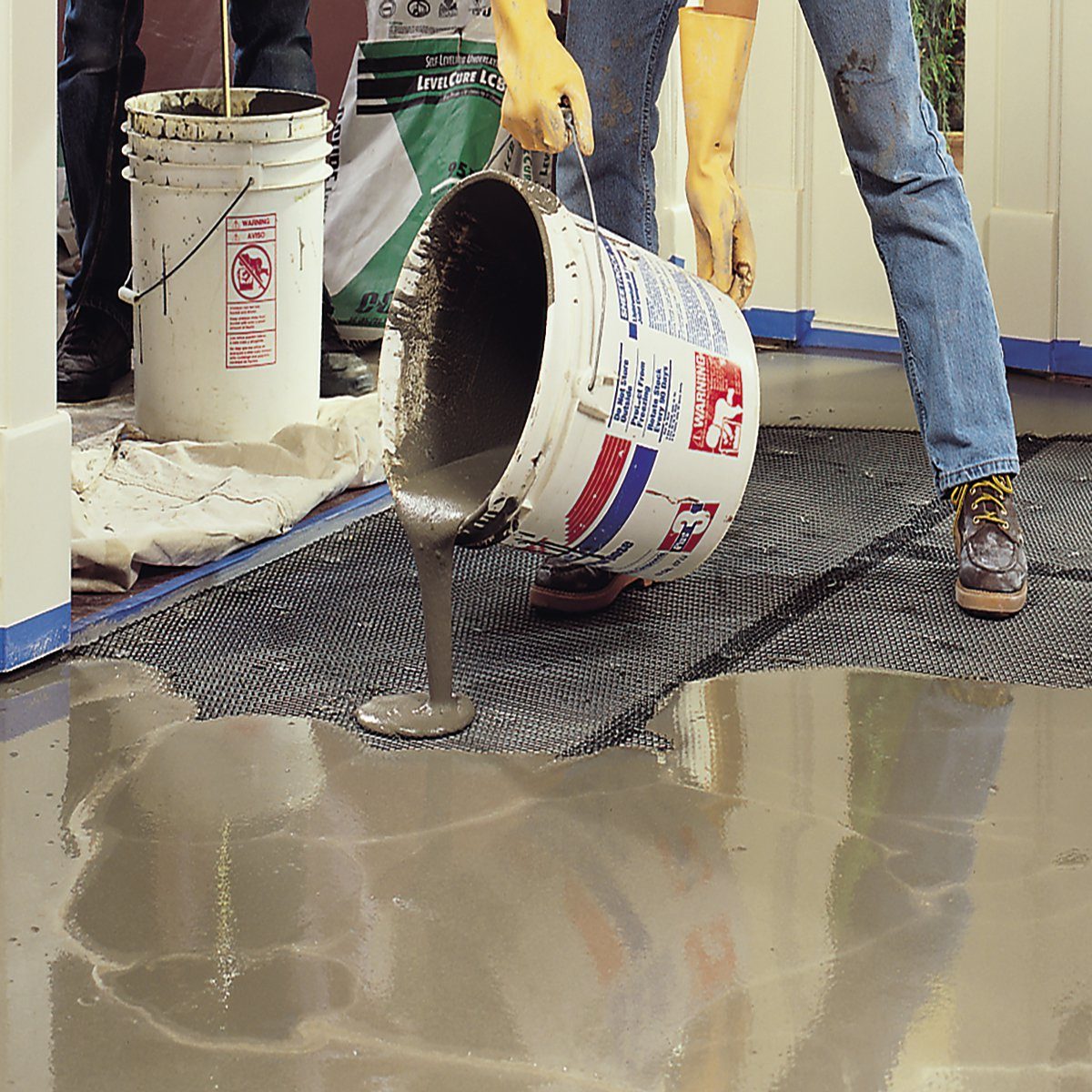 A person in gloves pours wet concrete from a bucket onto a textured surface, with buckets and a protective tarp visible in the background.