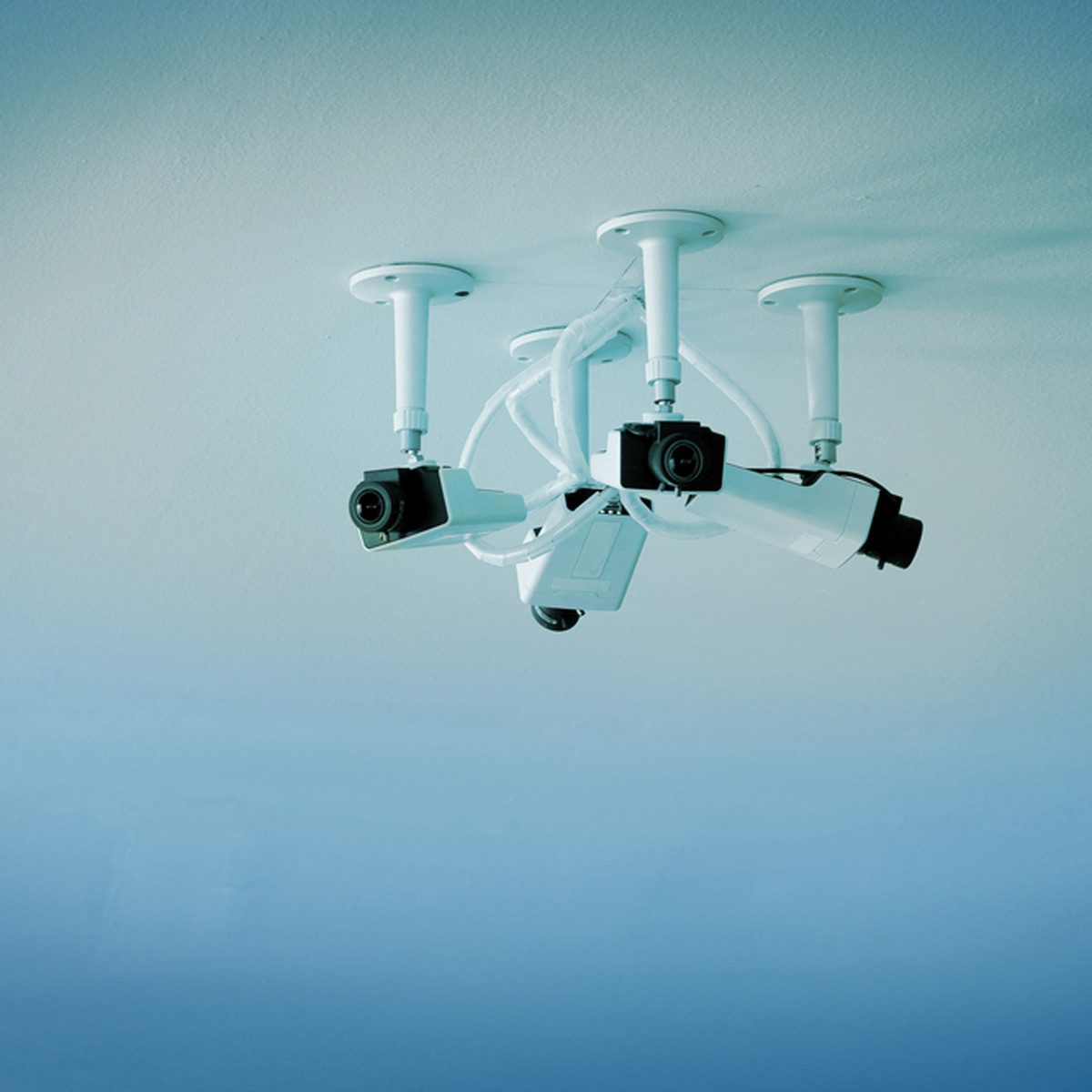 Four surveillance cameras are mounted on the ceiling, connected by cables, set against a smooth, light blue background.