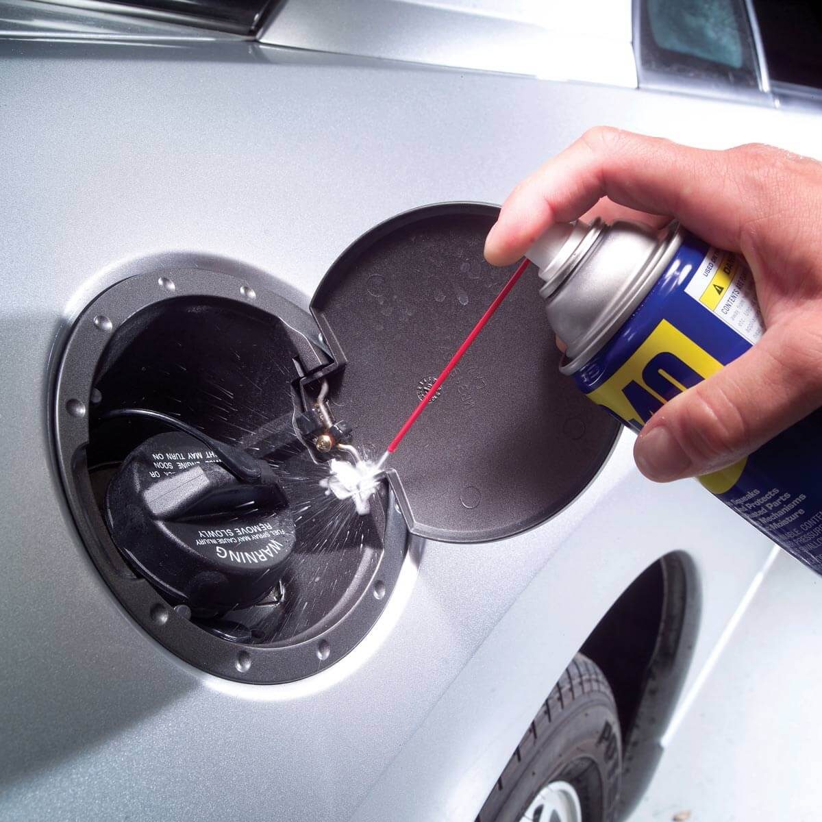 A hand sprays lubricant into a car's fuel cap area, potentially maintaining or cleaning a component, with a silver vehicle backdrop and visible tire.