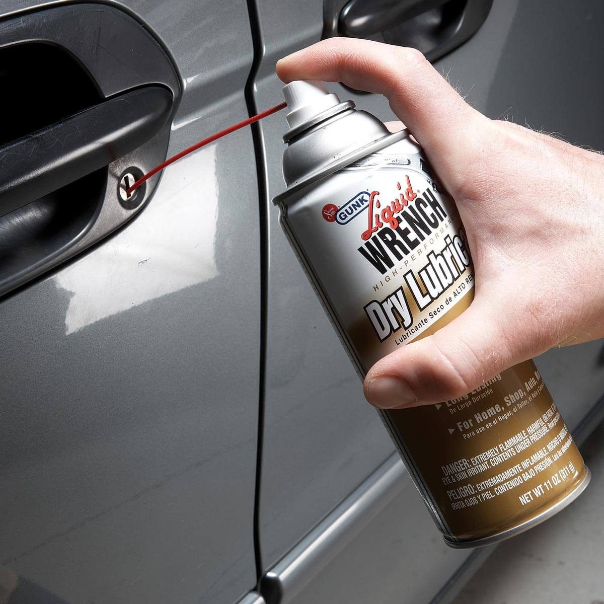 A hand holds a spray can, applying lubricant into a car door lock, positioned beside a sleek silver vehicle.