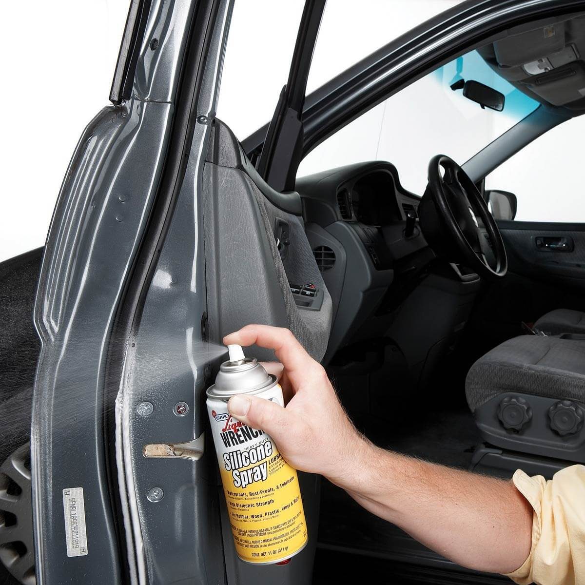 A hand sprays silicone lubricant onto the car door frame, enhancing the vehicle's functionality in an indoor setting with gray upholstery and a white backdrop.