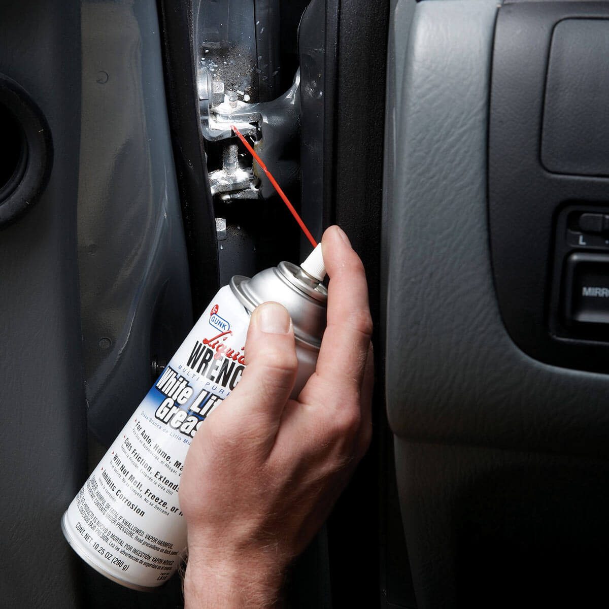 A hand holds a spray can, applying lubricant to a metal hinge within a vehicle's door frame, aimed at reducing friction and improving movement.