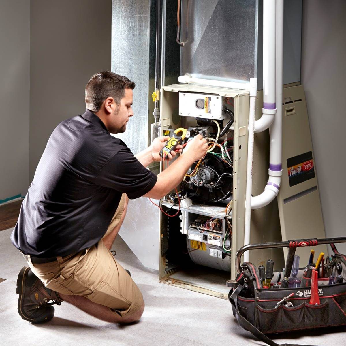 A technician kneels, examining wires inside a furnace, using a multimeter, in a dimly lit indoor space with a toolbox nearby.