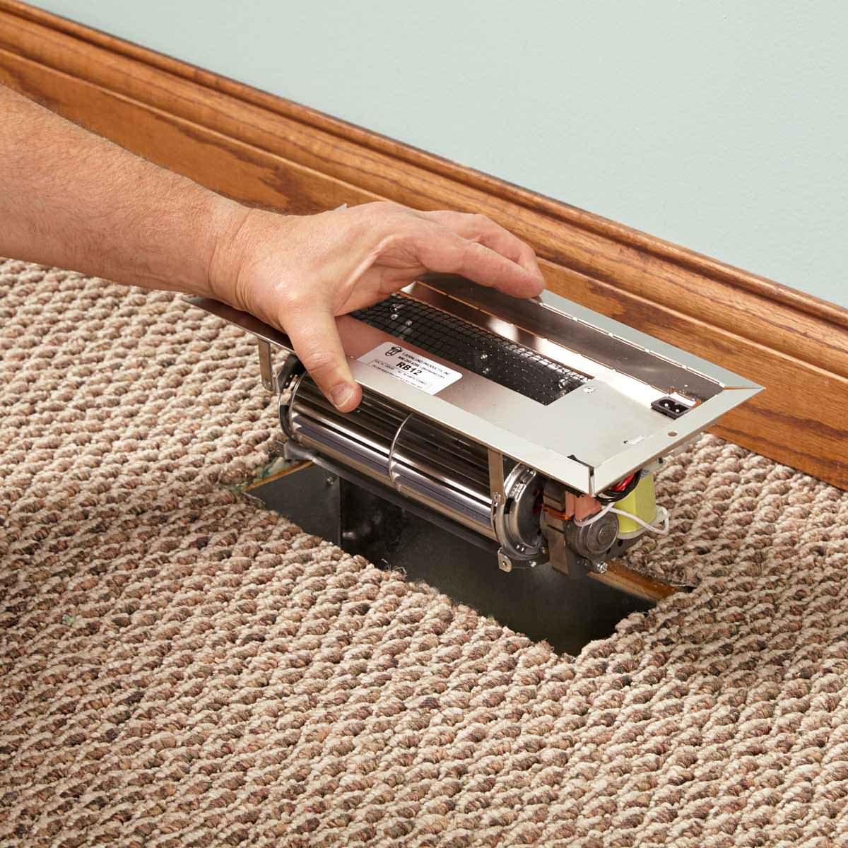 A hand adjusts a register vent on carpeted flooring near a wooden baseboard, revealing mechanical components underneath for air circulation.