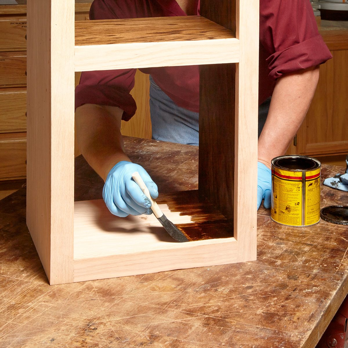 A person wearing gloves applies wood stain with a brush inside a wooden shelf structure, on a workbench surrounded by tools and materials.