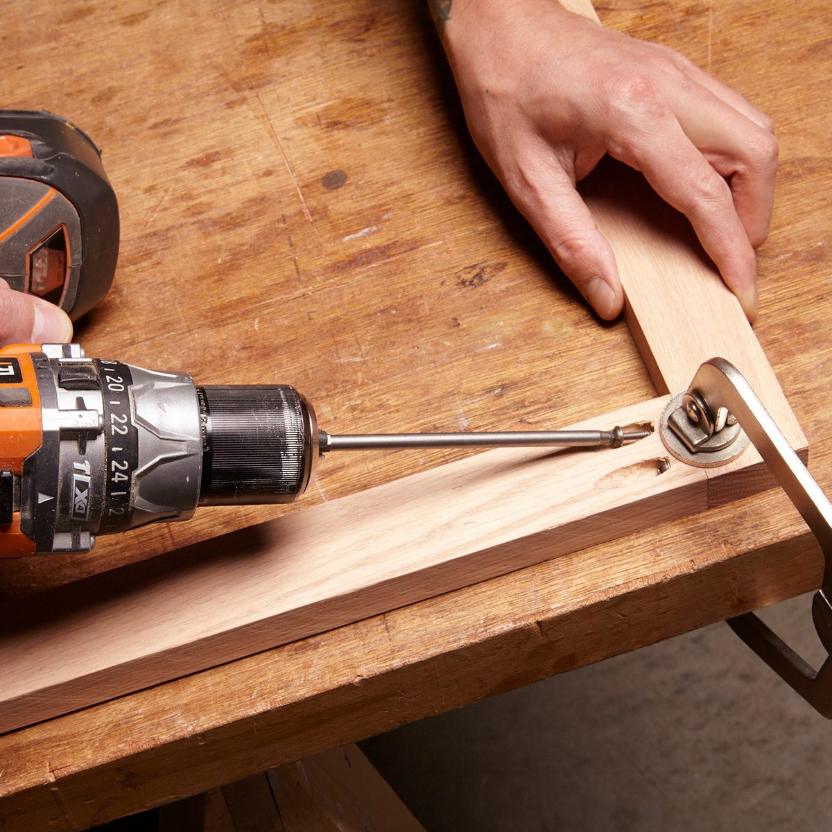 A drill is driving a screw into wooden pieces on a workbench, while a hand holds a clamp nearby for stability.