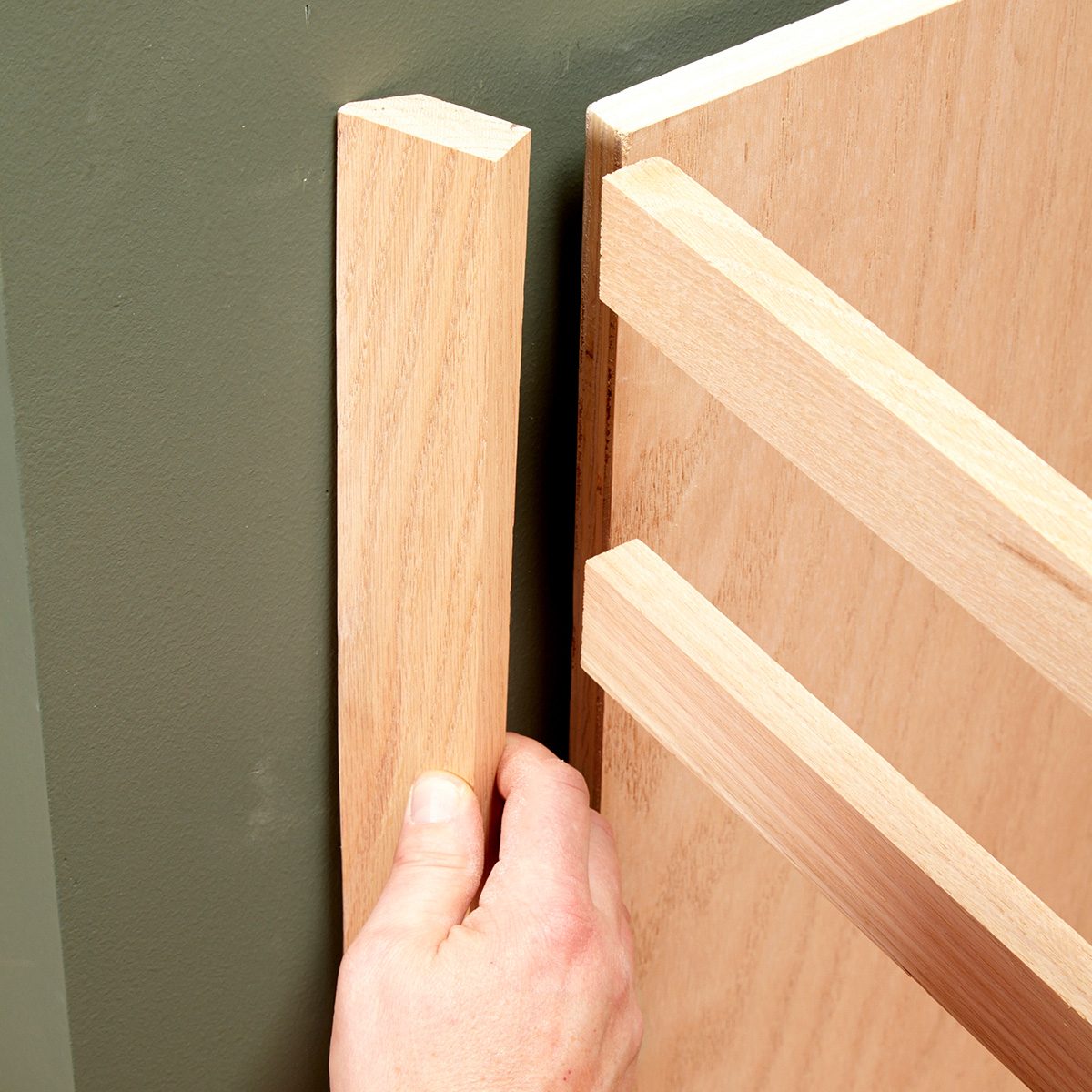 A hand is positioning a wooden piece adjacent to a panel, against a green wall, for assembly or construction purposes.