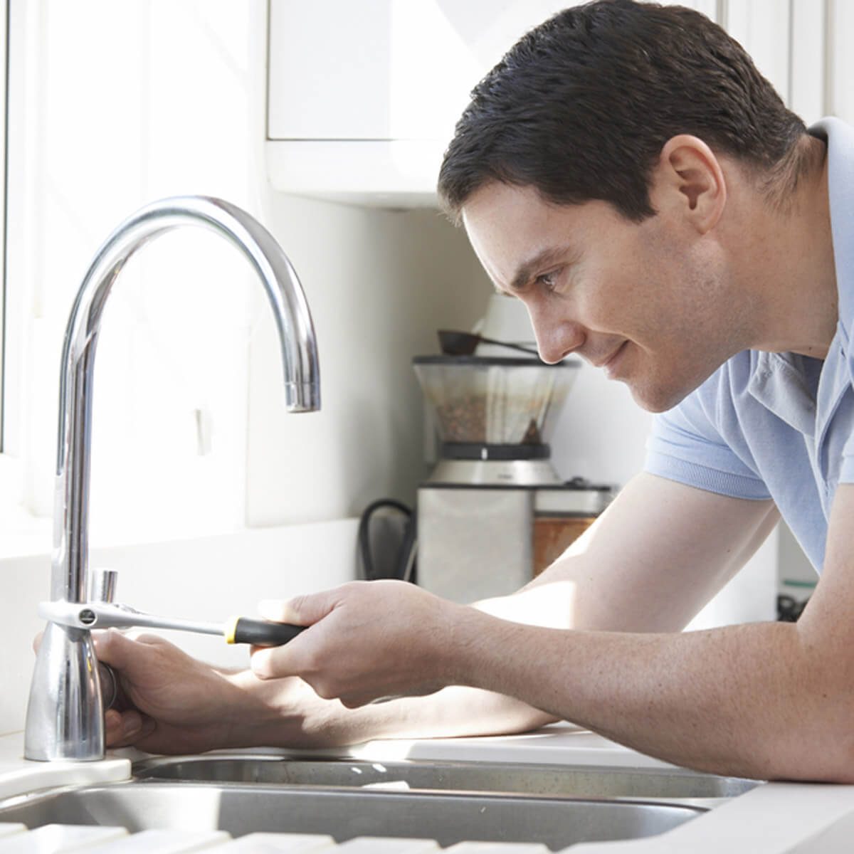 A man repairs a kitchen faucet, using a wrench, focused on the task. Natural light streams in, illuminating the clean, modern kitchen surroundings.