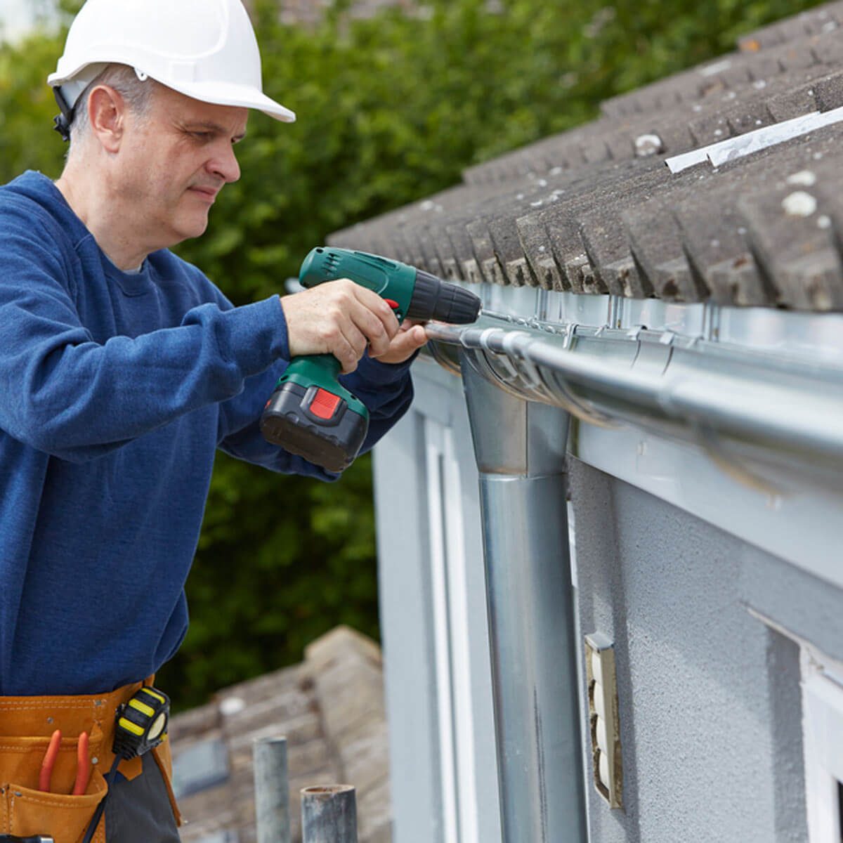 A man in a hard hat uses a power drill to attach a metal gutter to a roof, surrounded by greenery and construction materials.