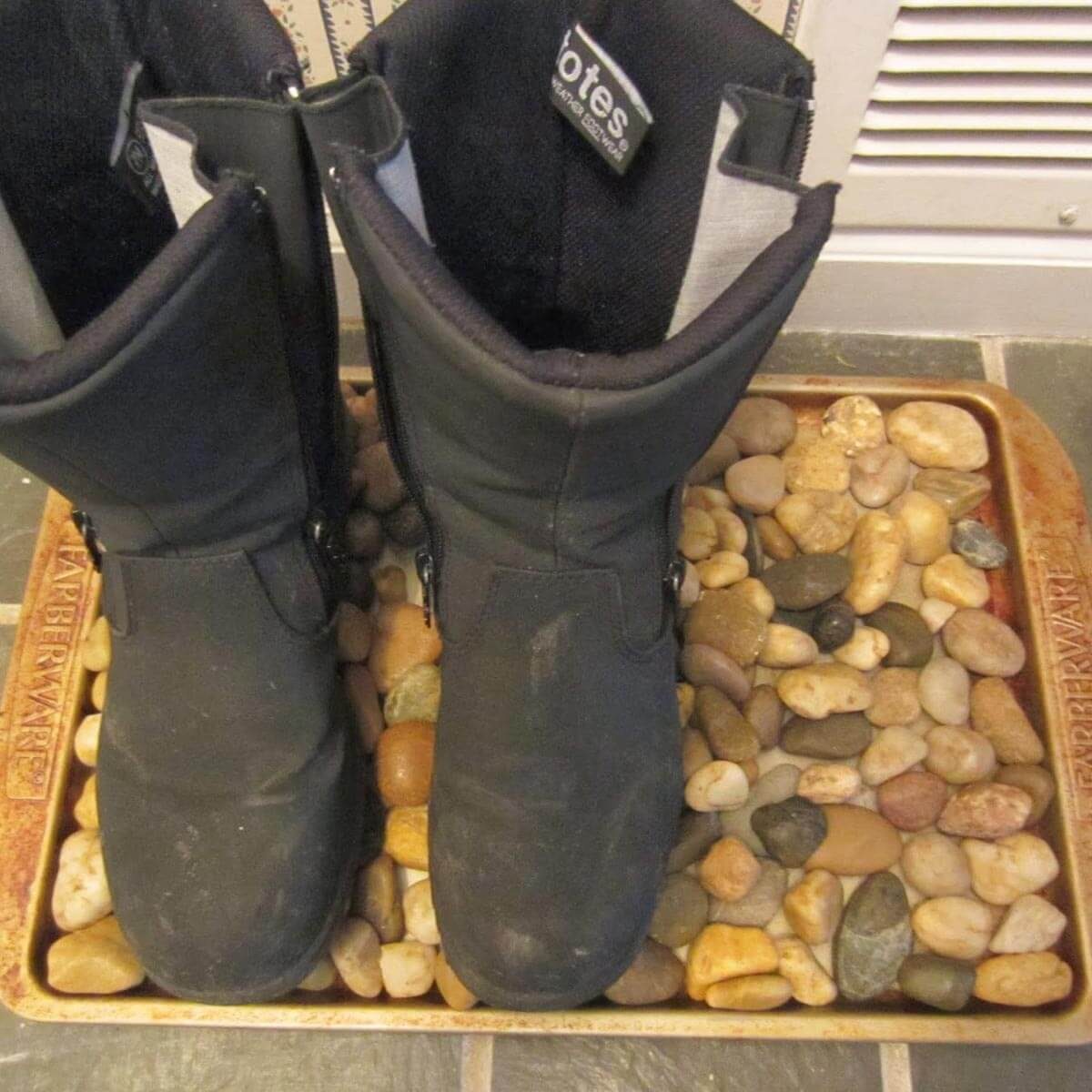 Black boots rest on a tray filled with assorted pebbles, situated on a tiled floor, indicating a mudroom or entryway setting.