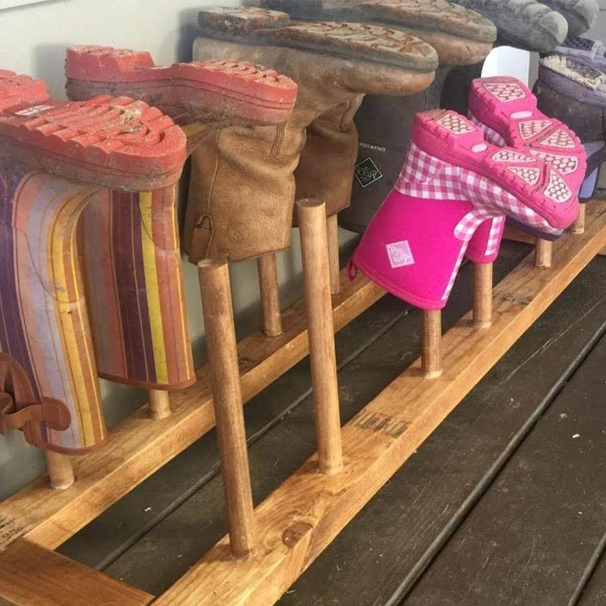 A shoe rack displays various pairs of colorful footwear on wooden supports, with boots and a pink pair prominently arranged in an indoor setting.