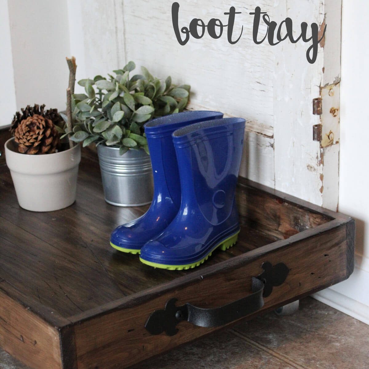 Blue rubber boots sit on a wooden boot tray, accompanied by potted plants and pinecones, in a cozy entryway. The text above reads "boot tray."