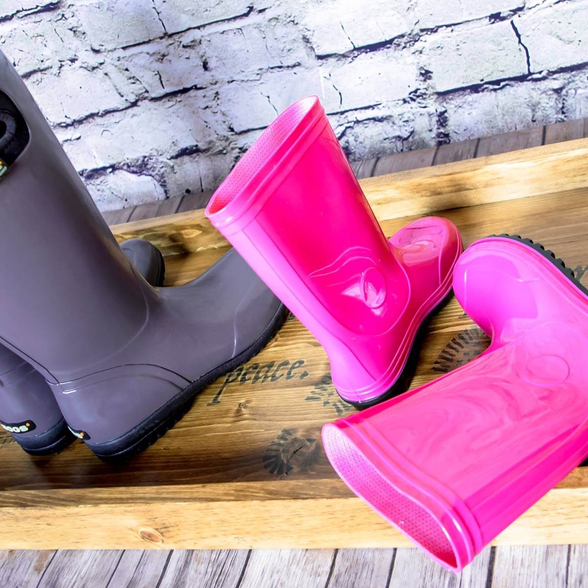 Two pairs of colorful rubber boots, one gray and one pink, are placed on a wooden tray with a decorative background featuring a brick wall.