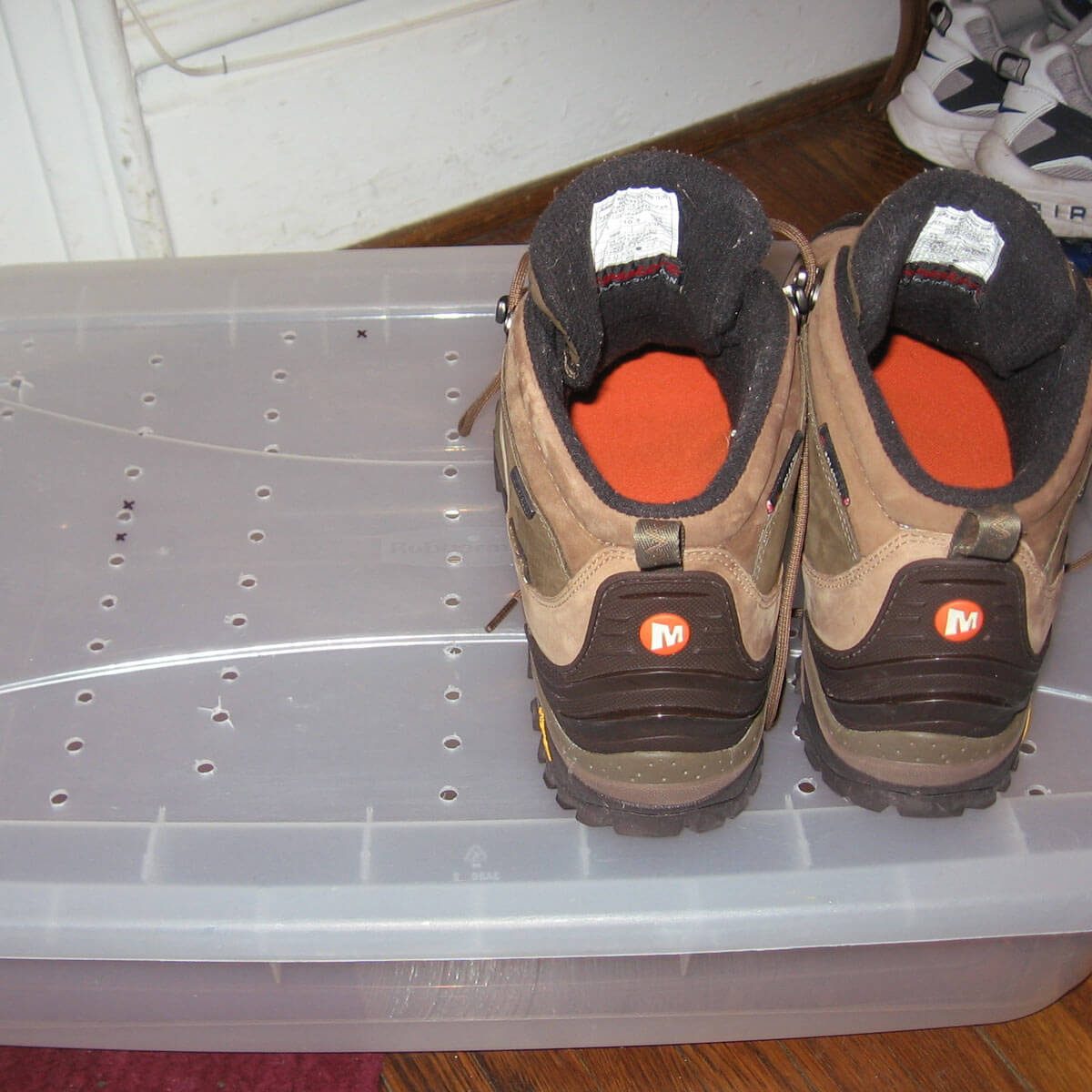 Brown hiking boots rest on a clear, perforated storage container placed on a wooden floor, surrounded by a white wall and a pair of sneakers in the background.