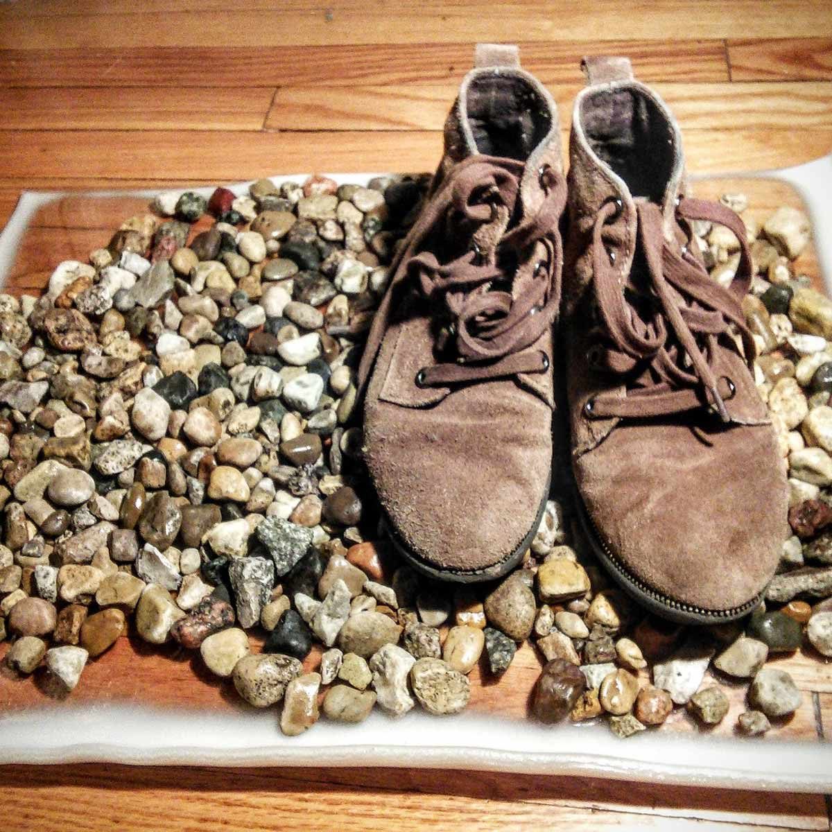Brown shoes rest on a diverse collection of small stones, surrounded by a clear material, with a wooden floor visible in the background.