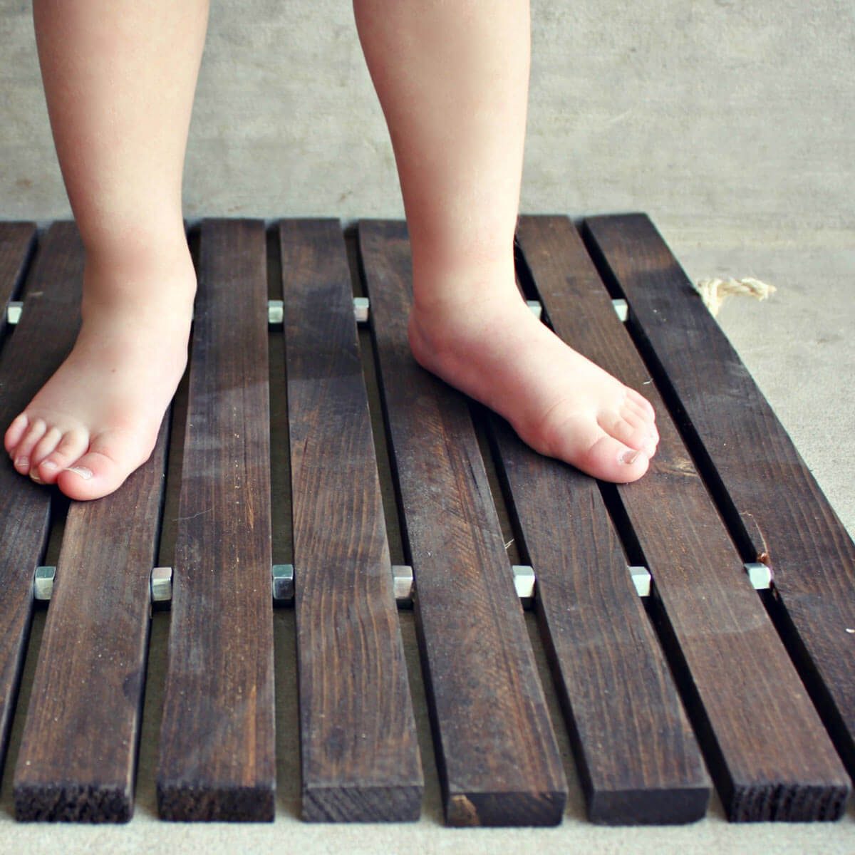 A bare child’s feet stand on a wooden slatted mat, positioned on a light, textured surface, suggesting a relaxed indoor environment.