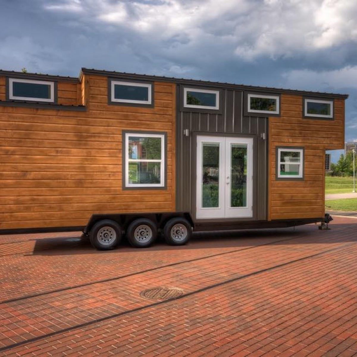A mobile tiny house, featuring wooden and metal exteriors, sits on a brick surface under a partly cloudy sky, accessible by double front doors.