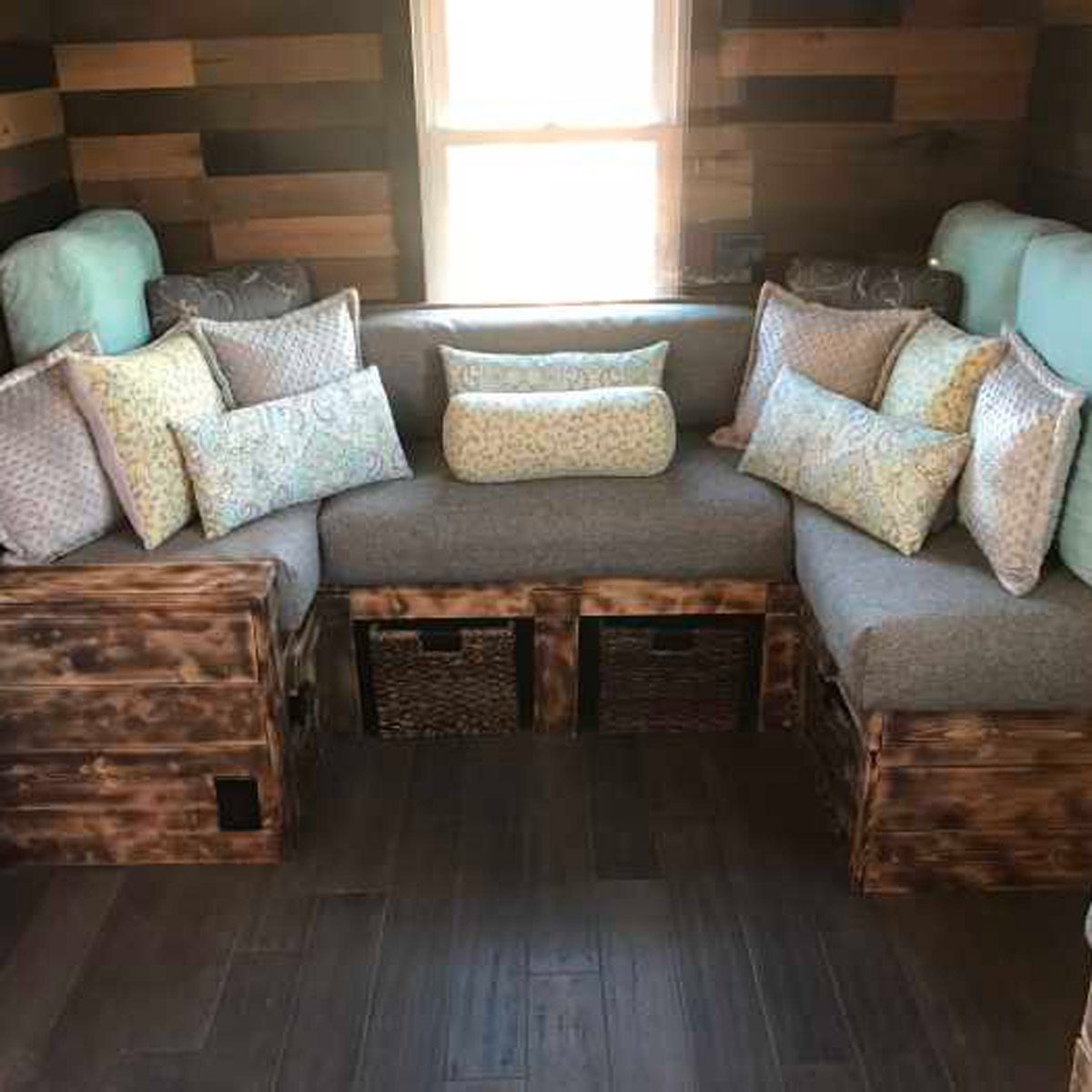A wooden corner couch with various patterned pillows rests against a wall, illuminated by natural light from a nearby window, on a wooden floor.