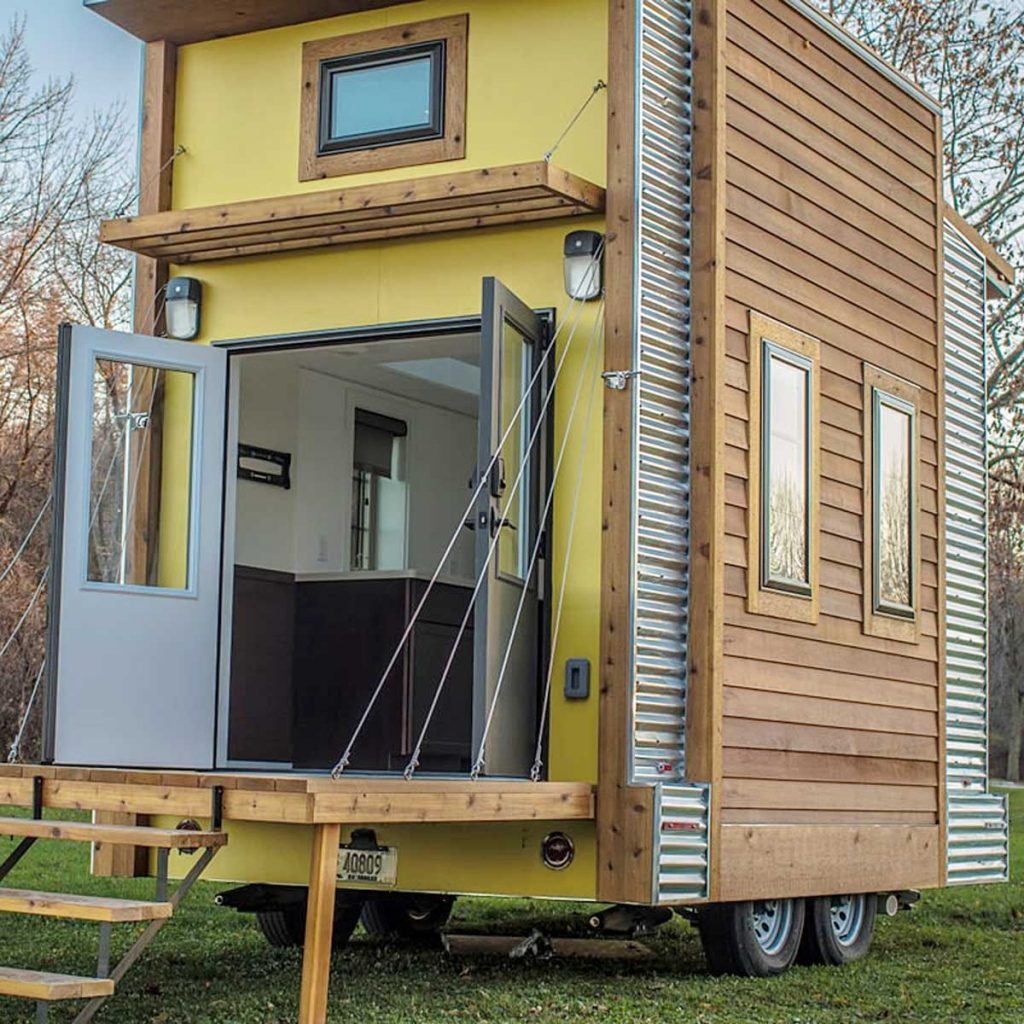 A yellow and wood tiny house, featuring open doors and a staircase, is situated on grass, surrounded by bare trees in a rural setting.