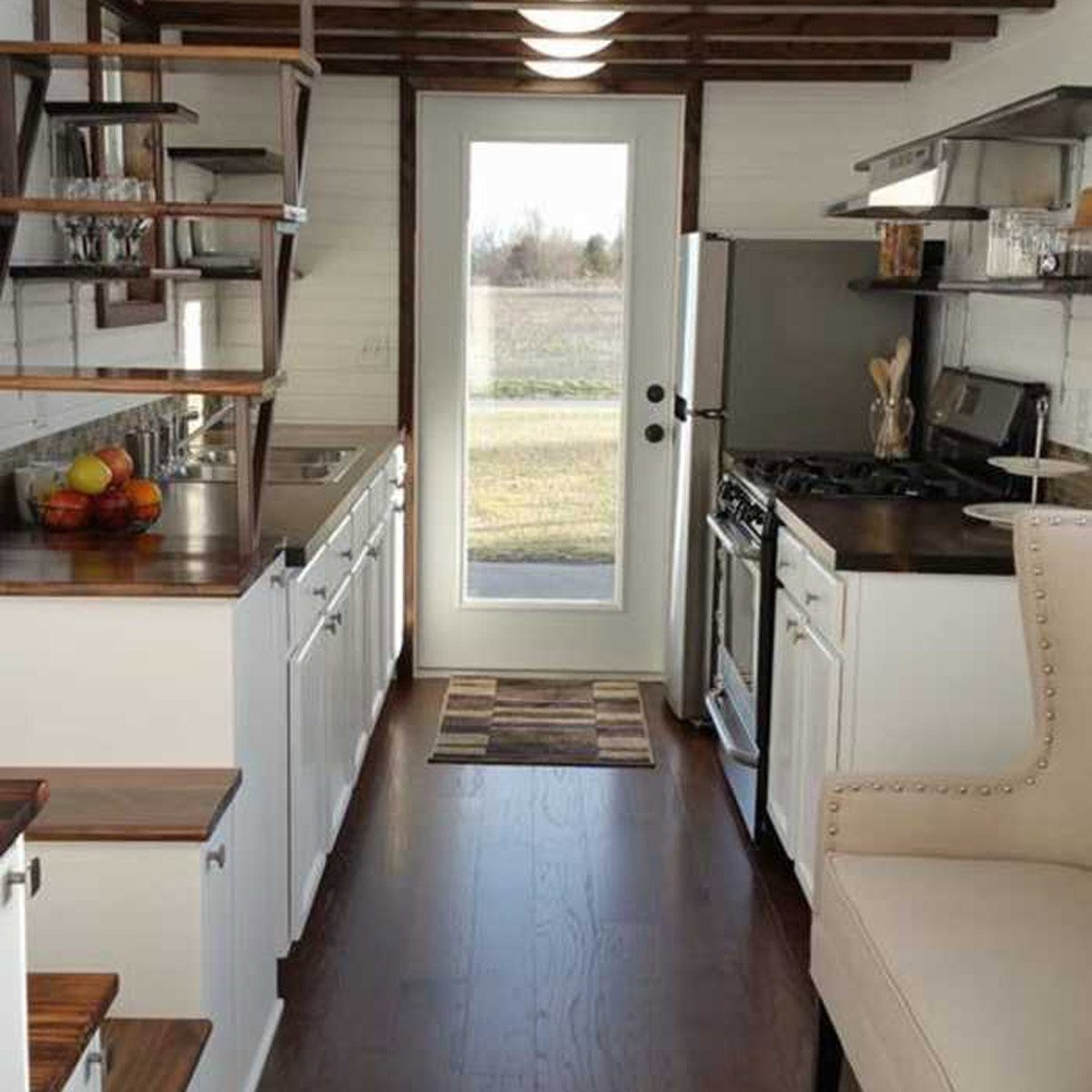 A modern kitchen features a counter with fruit, appliances, and a door leading to an outdoor view. Light floods in through a large window.