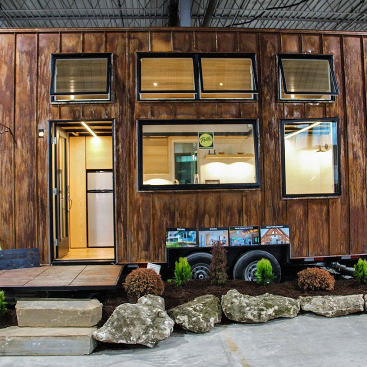A wooden tiny house features large windows and a door, positioned on a wheeled base. It is surrounded by landscaping elements like rocks and plants indoors.
