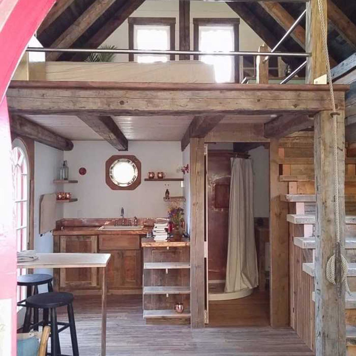A rustic kitchen features wooden cabinets and a circular window, with shelves displaying decor. A small staircase leads to a lofted sleeping area.