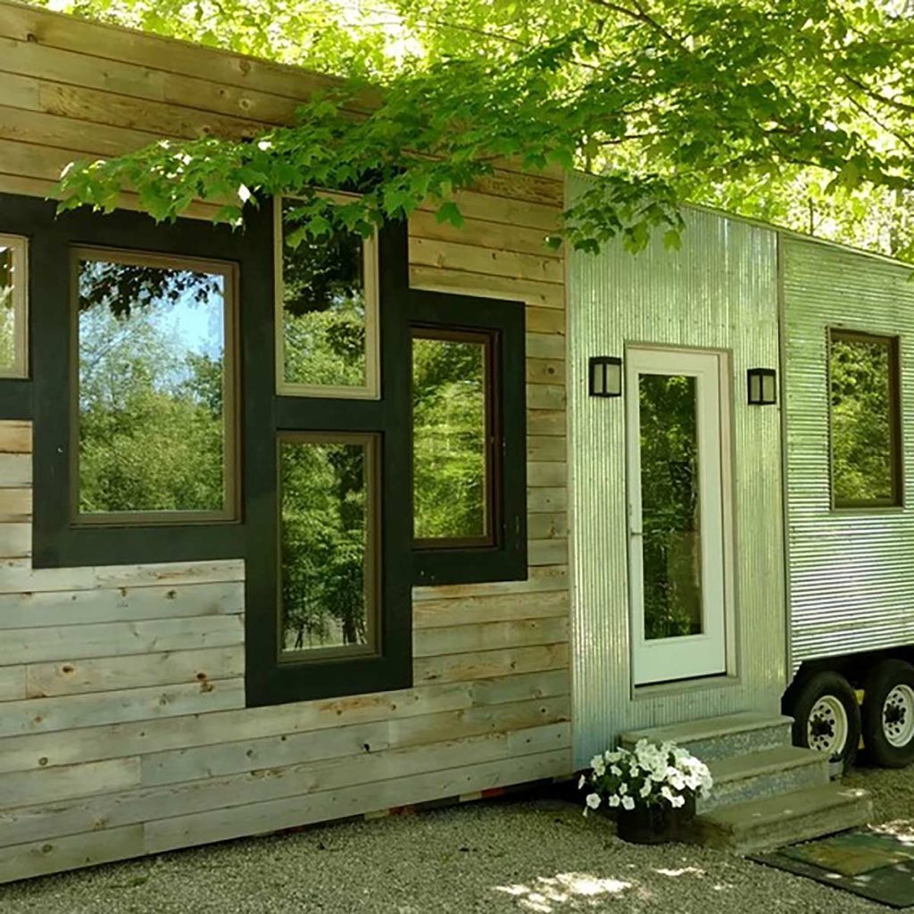 A modern tiny house features wooden and metal siding, large windows, and a front door, set amongst greenery and gravel landscaping with potted flowers.