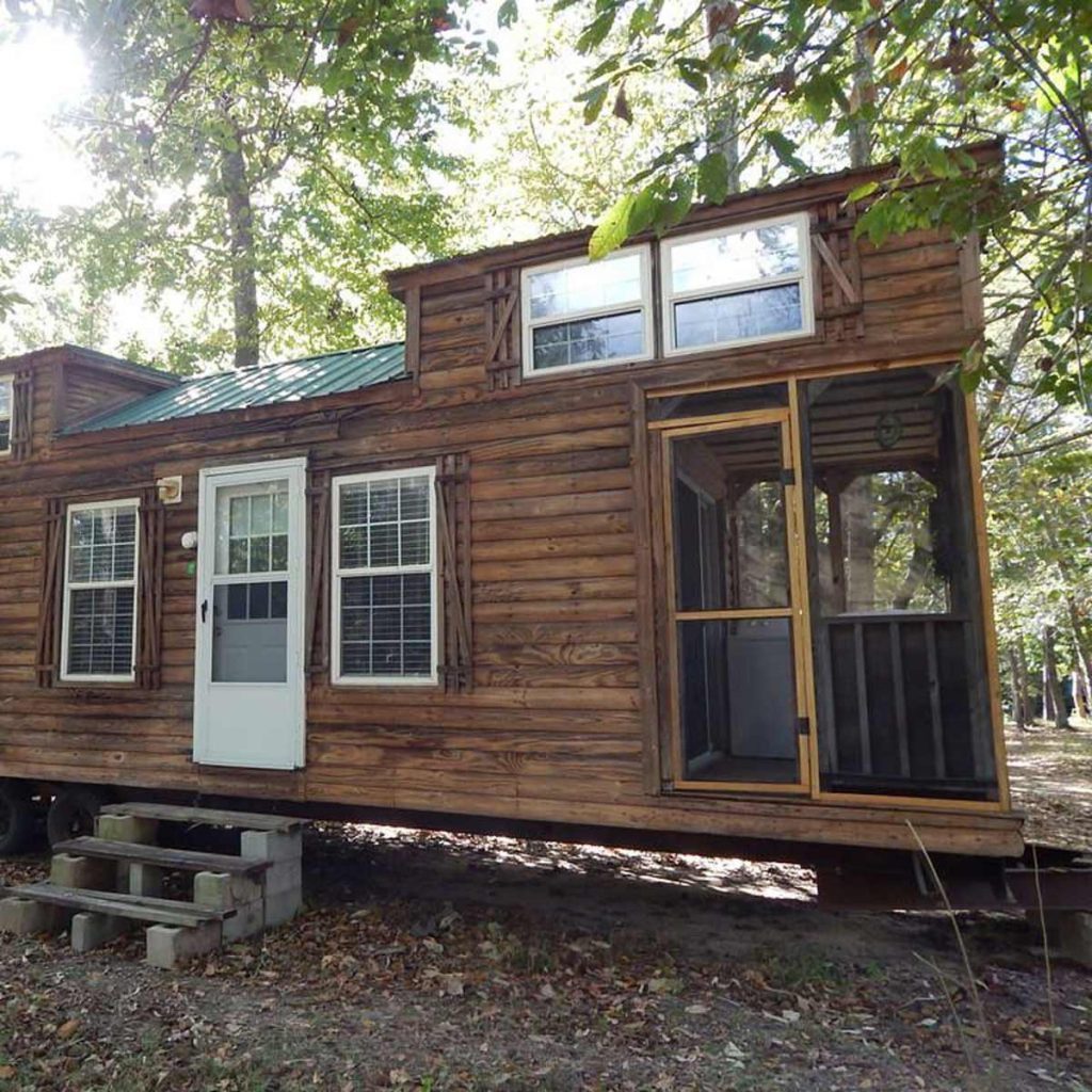 A wooden tiny house rests on blocks, featuring a front door and windows, situated in a forested area with sunlight filtering through the trees.