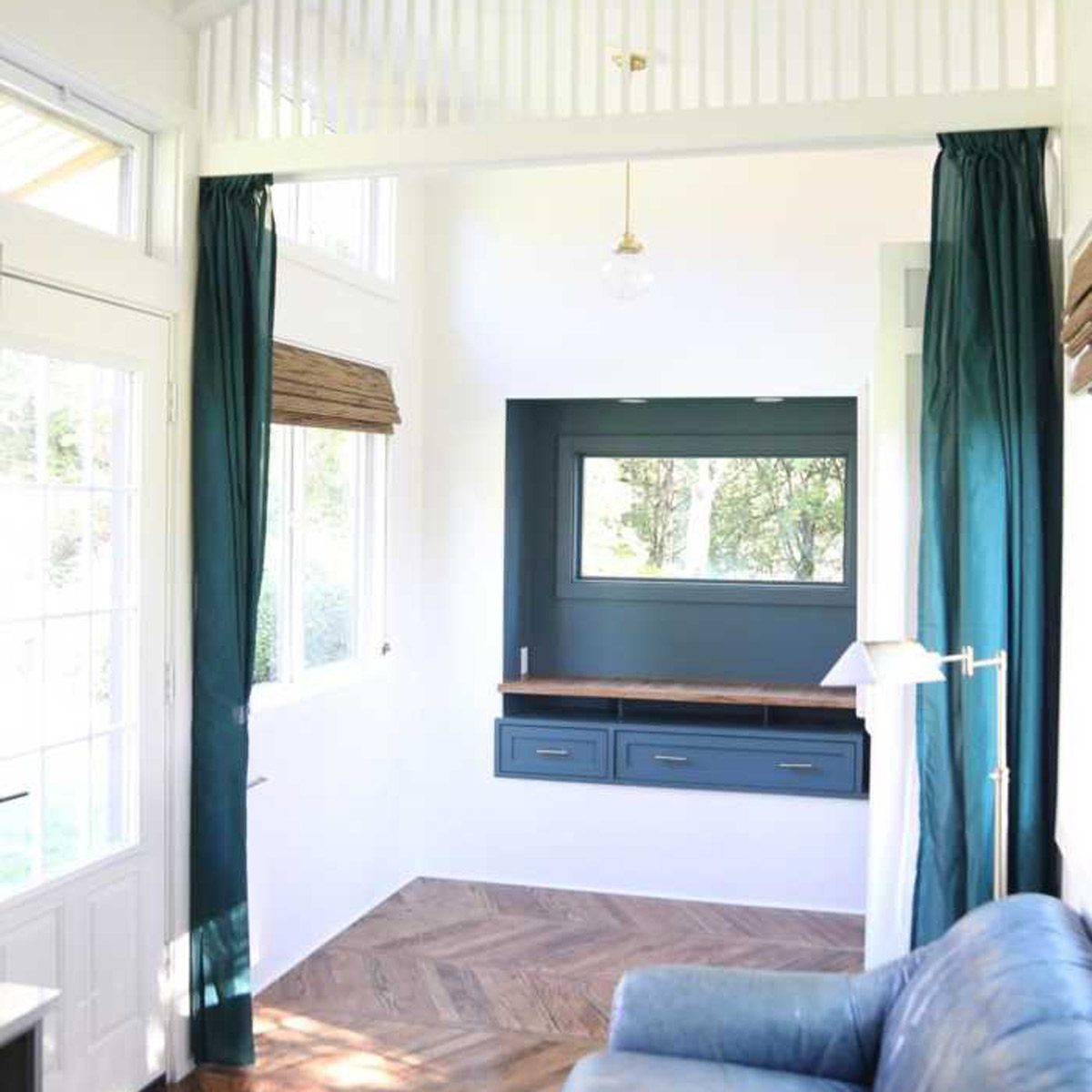 A cozy room features a blue sofa, wooden desk with drawers, and green curtains, illuminated by a pendant light, surrounded by large windows.