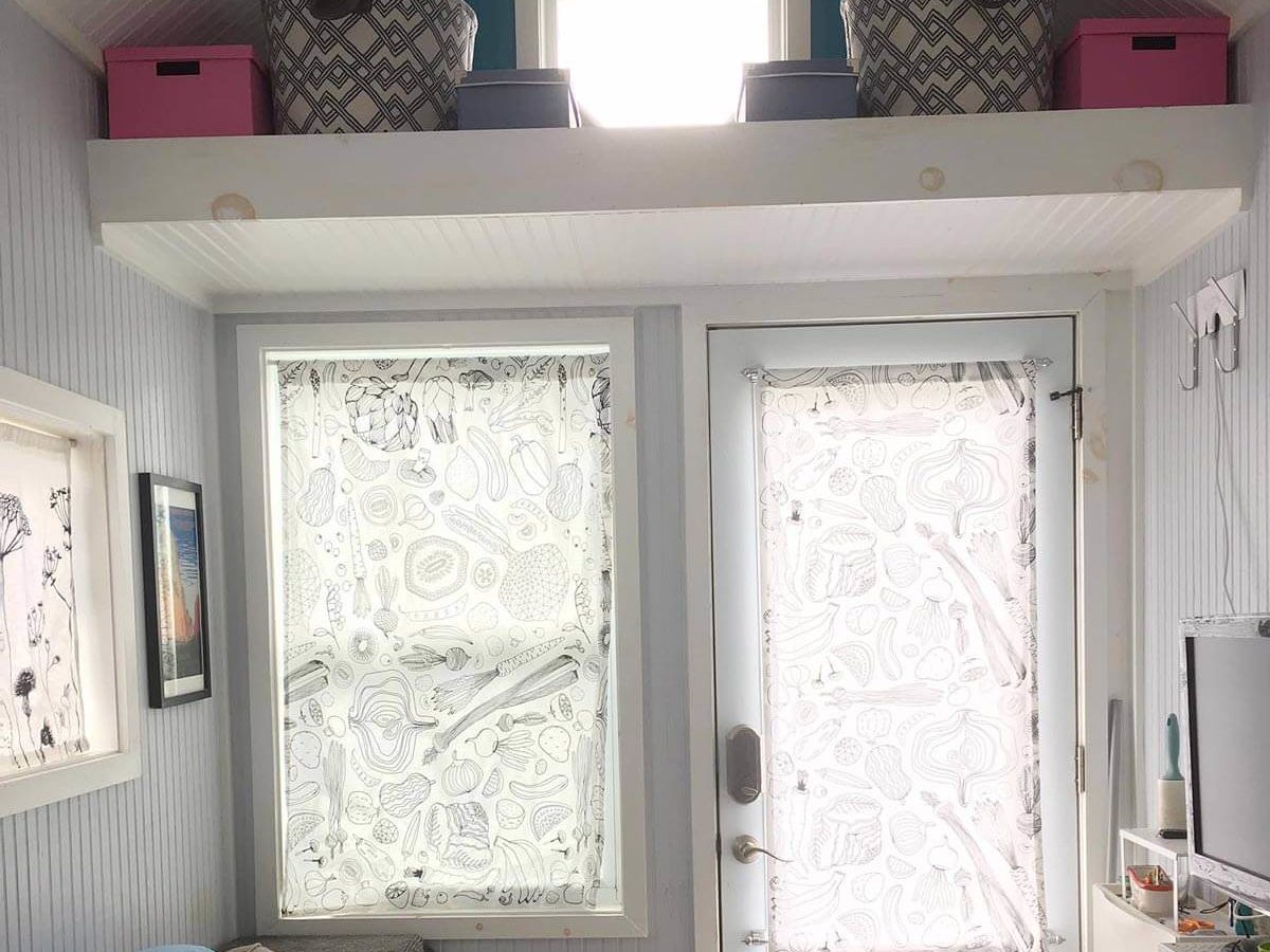 A living space features a door and patterned curtains. Shelves hold storage baskets above, illuminated by natural light. A couch sits in the corner.