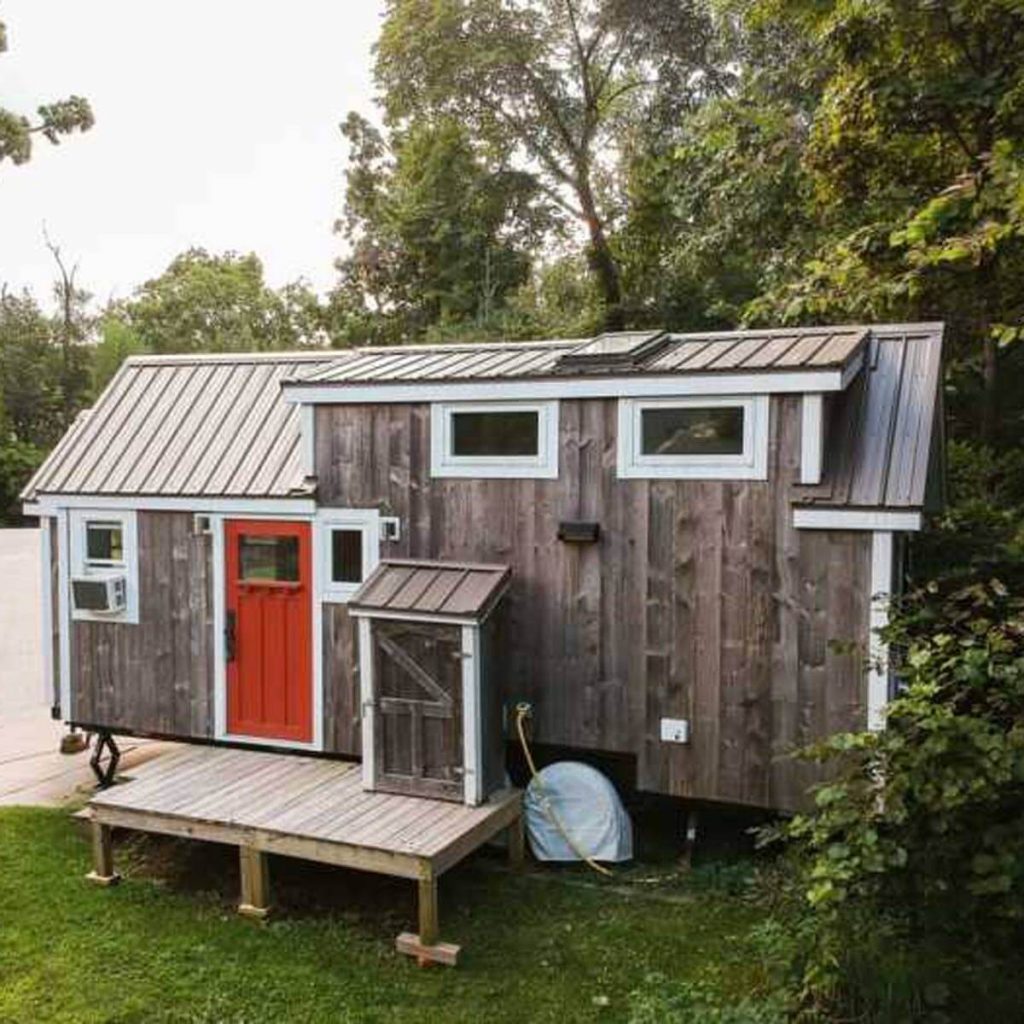 A small, wooden house with a bright red door sits on a wooden deck, surrounded by lush green trees and grass.