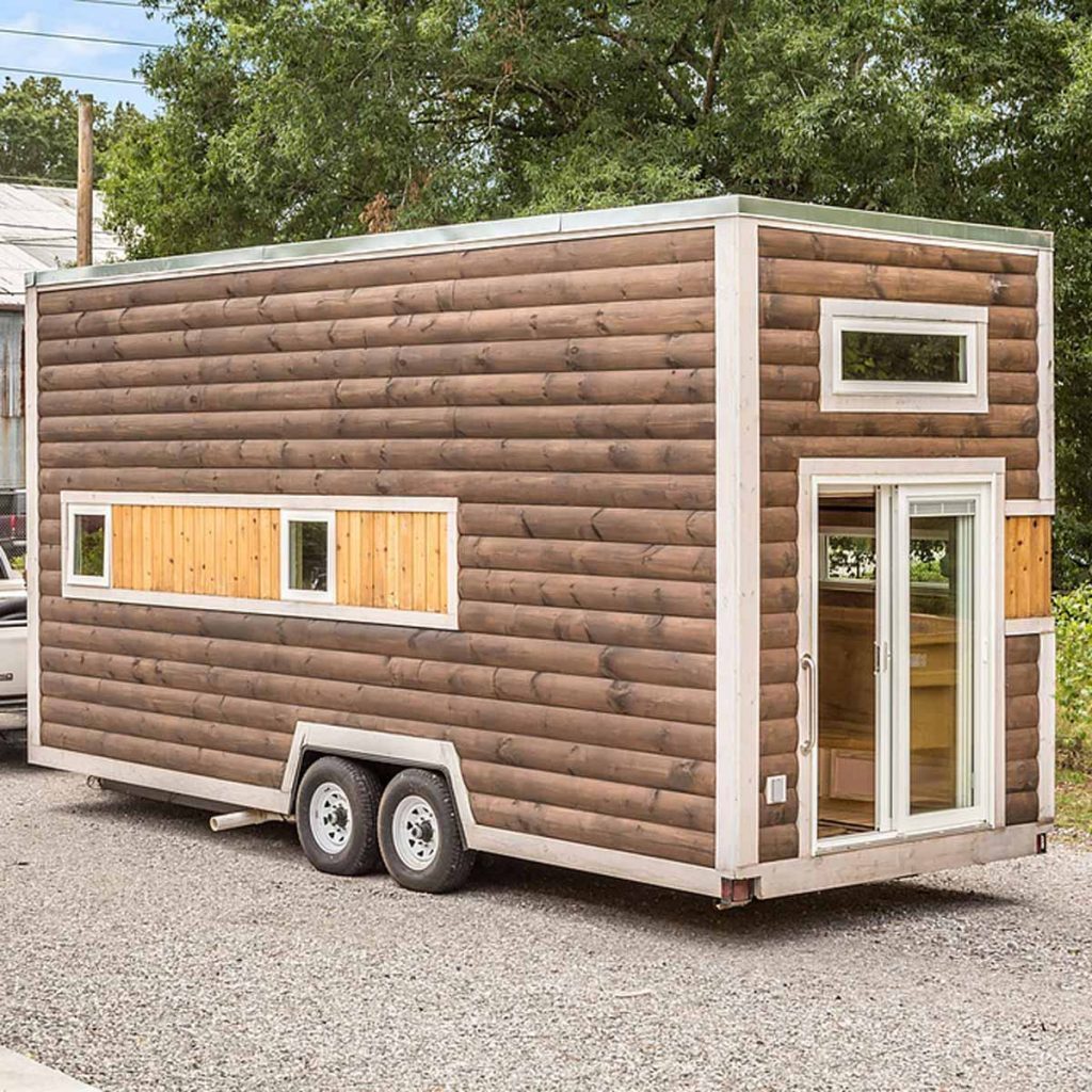 A wooden tiny house on wheels stands parked on gravel, featuring multiple windows and a door, amidst greenery and a nearby structure.