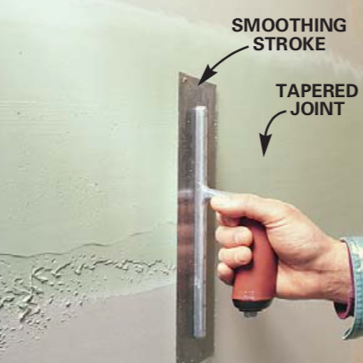 A hand holds a smoothing tool against a textured wall, applying a smoothing stroke to create a tapered joint in fresh drywall.