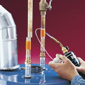 A person holds a torch, applying heat to copper pipes connected to a blue tank, with shiny metal components against a red background.